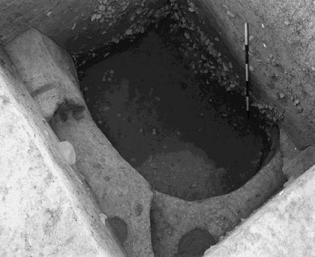 An excavation photograph of a deep oval pit dug into yellow karewa loess, with a measuring scale visible against the pit wall