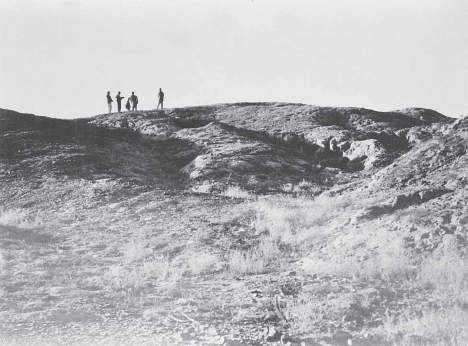 A long Harappan mound rising out of a flat, dry desert plain, with surveyors silhouetted on top